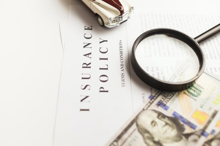 featured image that contains a white document labeled insurance policy, a $100 bill, and a magnifiying glass